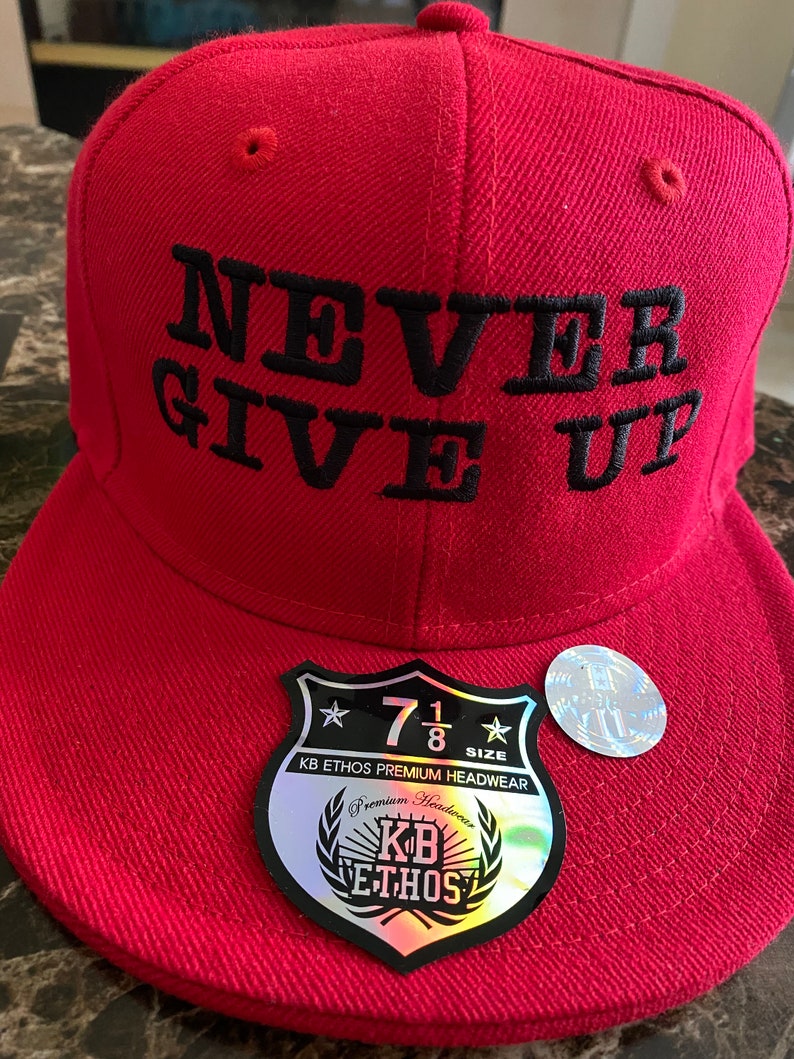 Puede incluir: Gorra de b&eacute;isbol roja con un mensaje bordado en negro que dice "NEVER GIVE UP". La gorra tiene una etiqueta negra y plateada con el texto "KB ETHOS PREMIUM HEADWEAR" y "7 1/8 SIZE".