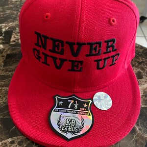 Puede incluir: Gorra de b&eacute;isbol roja con un mensaje bordado en negro que dice "NEVER GIVE UP". La gorra tiene una etiqueta en blanco y negro con el texto "KB ETHOS PREMIUM HEADWEAR" y una estrella con el n&uacute;mero "7 1/8" dentro.