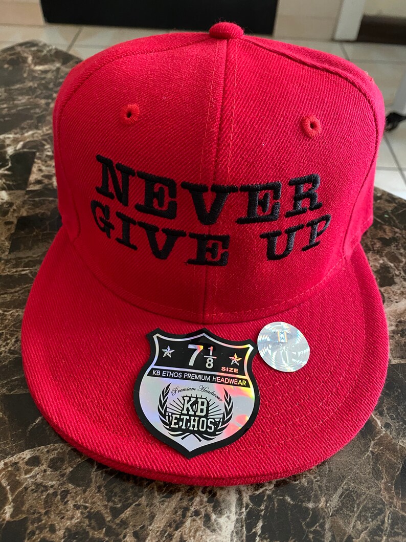 Puede incluir: Gorra de b&eacute;isbol roja con un mensaje bordado en negro que dice "NEVER GIVE UP". La gorra tiene una etiqueta en blanco y negro con el texto "KB ETHOS PREMIUM HEADWEAR" y una estrella con el n&uacute;mero "7 1/8" dentro.