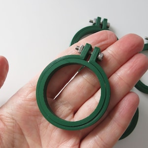 May include: Three green plastic embroidery hoops with metal screw closures. The hoops are different sizes and are held in a person's hand.