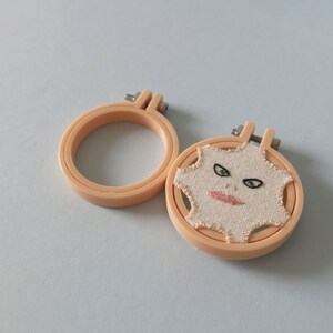 May include: Two embroidery hoops, one with a beige fabric featuring an embroidered face with green eyes and pink lips, and the other with a plain beige fabric.