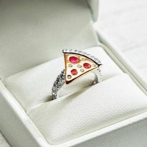Può includere: Un anello color argento con un elemento centrale a forma di fetta di pizza, caratterizzato da una crosta dorata, tre gemme rotonde rosse e piccole pietre trasparenti incastonate lungo la fascia e sul bordo superiore della fetta, in una scatola bianca.