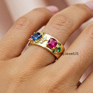Puede incluir: Anillo dorado con piedras preciosas azules, rojas y verdes. El diseño del anillo incluye una piedra azul en forma de corazón, una piedra roja cuadrada y una pequeña piedra verde. El anillo está en una mano.