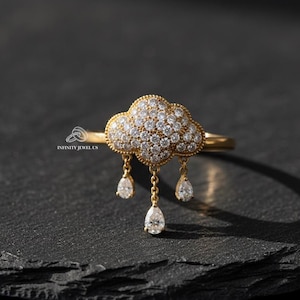 Könnte beinhalten: Ein goldener Ring mit einem Wolkenmotiv, besetzt mit kleinen, klaren Edelsteinen. Drei tropfenförmige Edelsteine hängen darunter. Der Ring ist auf einer dunklen, strukturierten Oberfläche platziert.