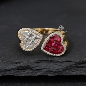 May include: A gold ring featuring two heart-shaped sections. One heart is filled with white stones, the other with red stones. Both hearts are outlined with small, clear stones. The ring is set against a dark background.