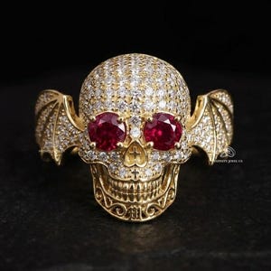 May include: A gold ring shaped like a skull with bat-like wings on the sides. The skull is covered in small clear pavé stones and features two round, deep red rubies as eyes. The metal surface has intricate engraved details around the jawline.