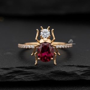 May include: A unique gold ring featuring a ruby-colored oval gemstone at the center, shaped like a beetle. The ring has a small diamond on top of the beetle's head and small diamonds along the band. The ring is set against a black background.