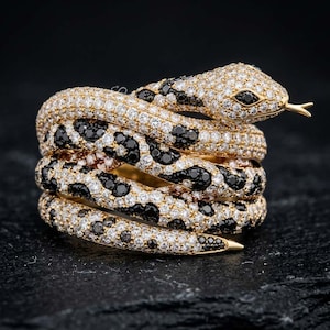 May include: A gold ring shaped like a coiled snake, featuring a pavé setting of numerous small, clear white stones and clusters of black stones arranged in a spotted pattern along the body, with two black stones forming the snake's eyes.