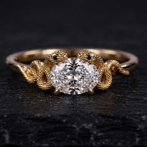 Puede incluir: Un anillo de oro con dos figuras de serpiente texturizadas frente a frente, cuyas cabezas se unen para sostener una piedra preciosa transparente de corte ovalado en el centro. Las serpientes tienen pequeñas piedras azul oscuro como ojos.