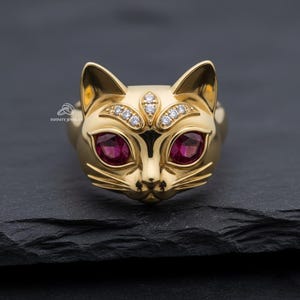May include: A gold cat ring with ruby eyes and diamond detailing. The ring has a detailed cat face design, including whiskers and pointed ears. The ring is set against a dark background.
