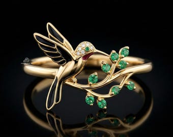May include: A gold ring featuring a hummingbird design. The bird has black enamel wings, a red beak, and a body adorned with diamonds. It perches on a branch with green emerald leaves. The ring is set against a black background.