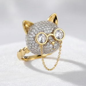 May include: A gold ring in the shape of a cat's head, encrusted with small, sparkling stones. The cat wears round, clear spectacles with a fine chain. The ring's band and ears are gold. A distinctive piece of jewellery.