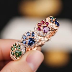 May include: Rose gold ring featuring paw print designs, each adorned with a variety of colorful gemstones. The ring showcases a central sapphire, surrounded by smaller gems in shades of green, blue, red, and purple, and accented with small white diamonds.
