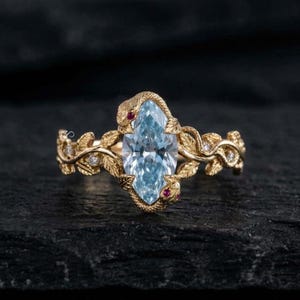 May include: A gold ring featuring a central marquise-cut light blue gemstone. The band is designed with textured leaf motifs and two small snakes with red gemstone eyes. Several tiny clear accent stones are embedded along the golden vine band.