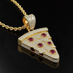 May include: A gold-colored pizza slice pendant necklace. The pendant is encrusted with small, clear gemstones and features red gemstone toppings. The necklace chain is also gold-colored and has a rope design.