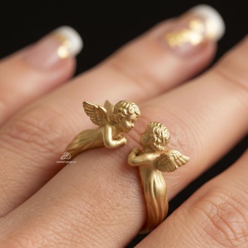 Puede incluir: Un anillo dorado con dos figuras de querubines alados. Un querub&iacute;n est&aacute; posicionado en el lateral del anillo, mientras que el otro est&aacute; erguido, creando un dise&ntilde;o &uacute;nico y detallado. El anillo se muestra en un dedo.
