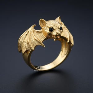 Może przedstawiać: Złoty pierścionek z motywem nietoperza. Nietoperz ma szczegółowe skrzydła i czarne oczy. Pierścionek wykonany jest z błyszczącego, metalicznego materiału i osadzony na ciemnym tle. Pierścionek to biżuteria.