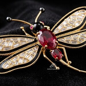 May include: A dragonfly brooch with a gold-tone frame, black enamel accents, and baguette-cut crystals. The brooch features a large oval ruby and smaller ruby accents. The dragonfly's eyes are black pearls.