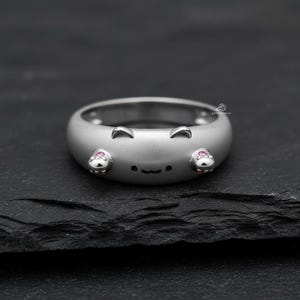 May include: A silver-toned ring with a matte finish featuring a stylized cat face design. The ring includes two small ears at the top, a simple black line for a mouth, and two small pink stones set into the sides to resemble cat paws.