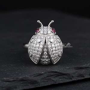 May include: A silver ladybug ring with a detailed design. The body is encrusted with small, clear gemstones, and the eyes are small red gems. The ring has antennae and is set against a dark background.