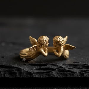 Puede incluir: Un anillo dorado con dos figuras de querubines con alas detalladas y cabello rizado. Los querubines se miran, creando un dise&ntilde;o &uacute;nico. El anillo est&aacute; sobre un fondo oscuro.