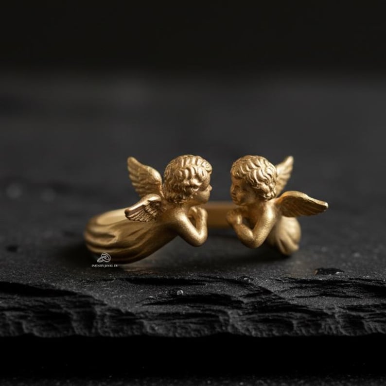 Puede incluir: Un anillo dorado con dos figuras de querubines con alas detalladas y cabello rizado. Los querubines se miran, creando un dise&ntilde;o &uacute;nico. El anillo est&aacute; sobre un fondo oscuro.