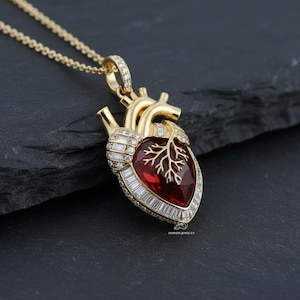 May include: A gold-colored pendant necklace featuring a heart-shaped pendant. The pendant has a red center with a tree-like design and is encrusted with clear gemstones. The necklace is displayed on a dark surface.