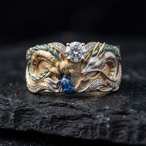 May include: A gold and silver ring featuring a central round brilliant-cut clear gemstone. The band depicts a sculpted figure with a ponytail in a blue top facing a long, winding dragon with green accents, surrounded by small embedded clear stones.