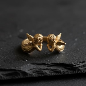 Puede incluir: Un anillo dorado con dos figuras de querubines con alas detalladas y cabello rizado. Los querubines se enfrentan, con las manos apoyando sus barbillas. El anillo est&aacute; sobre una superficie oscura y texturizada.