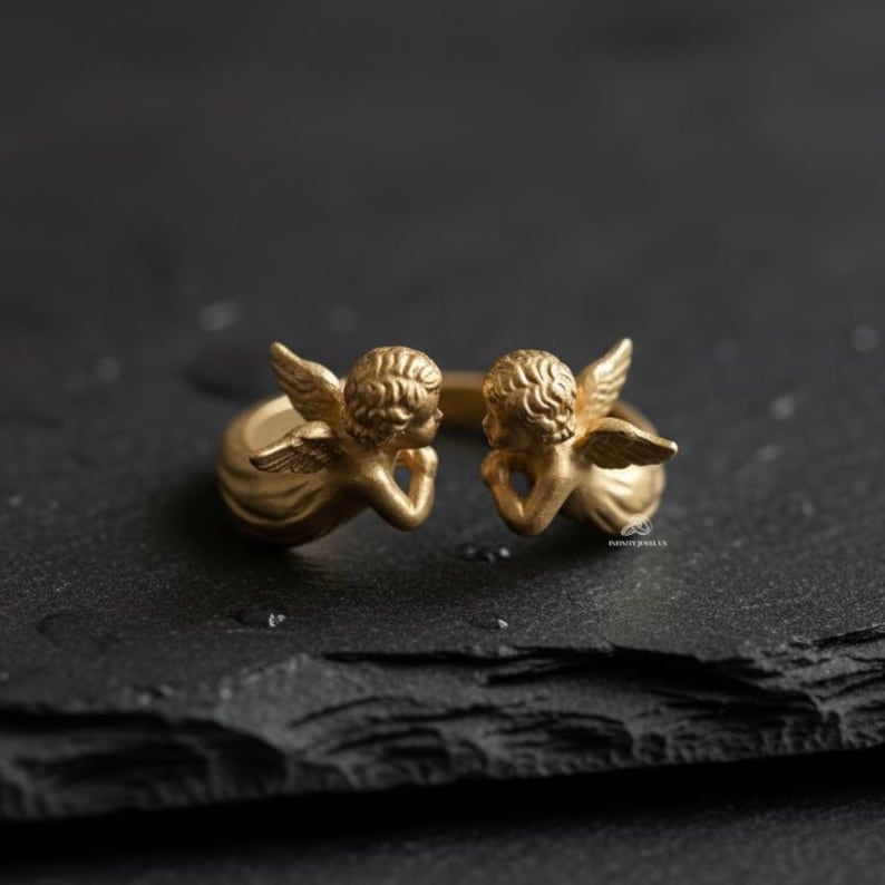 Puede incluir: Un anillo dorado con dos figuras de querubines con alas detalladas y cabello rizado. Los querubines se enfrentan, con las manos apoyando sus barbillas. El anillo est&aacute; sobre una superficie oscura y texturizada.