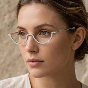 May include: A woman wearing half-rimmed eyeglasses. The frames are a light color and are encrusted with small, sparkling crystals. The lenses are a clear, curved shape. The background is a blurred, neutral tone.