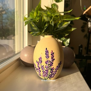 Könnte beinhalten: Eine gelbe Keramikvase mit handbemaltem Design aus lila Lavendelblüten. Die Vase steht auf einer Fensterbank mit einer grünen Pflanze im Hintergrund. Der Text "@laaroocheart" befindet sich am unteren Rand des Bildes.