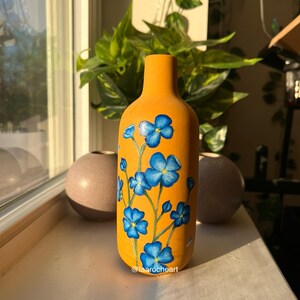 May include: A yellow ceramic vase with a hand-painted design of blue flowers. The vase is sitting on a windowsill with a plant in the background.