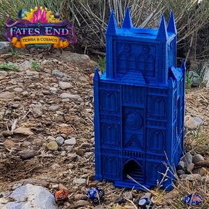May include: A blue 3D printed tower with a gothic design. The tower has a large arched doorway and several windows. The tower is sitting on a bed of gravel. There are several dice in the foreground, including a red and orange die, a blue die, and a black die.