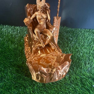 May include: A gold-colored 3D printed figurine of a woman with horns sitting on a throne. The throne is decorated with intricate details and the woman is holding a crystal in her hand. The figurine is sitting on a bed of green grass.
