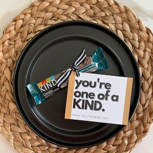 May include: A black and gold card with the text "you're one of a KIND. THANK YOU FOR BEING YOU!" and a KIND brand granola bar with a black and white ribbon tied around it.