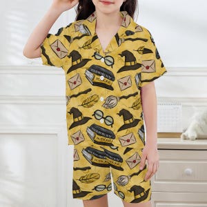 May include: Yellow pajama set with a repeating pattern of wizard hats, glasses, books, feathers, and envelopes. The text "Herbology Potions" is visible on the book. The set includes a short-sleeved button-up shirt and matching shorts.
