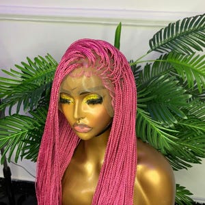 Può includere: Una parrucca rosa intrecciata su una testa di manichino. La parrucca presenta un frontale in pizzo e lunghe micro trecce. Il manichino ha la pelle dorata ed è ambientato su uno sfondo di fogliame verde. Il colore della parrucca è un rosa vibrante e accattivante.