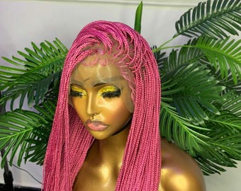 May include: A pink braided wig on a mannequin head. The wig features a lace front and long, micro braids. The mannequin has gold skin and is set against a backdrop of green foliage. The wig's color is a vibrant, eye-catching pink.