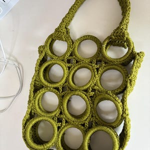 Puede incluir: Una bolsa de mano de crochet verde con un diseño circular. La bolsa tiene un solo asa y está hecha de hilo grueso.