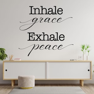 May include: A white wall with black text that reads "Inhale grace Exhale peace" with a decorative flourish under each phrase. A wooden shelf with a white surface sits below the text. A small, round, light gray stool sits in front of the shelf.