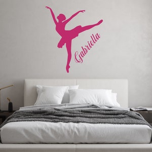 May include: Pink ballerina silhouette wall decal with the name Gabriella written in a cursive font. The decal is on a white wall above a white bed with a gray blanket.