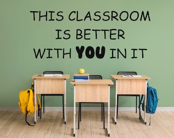 May include: A green wall with the text "THIS CLASSROOM IS BETTER WITH YOU IN IT" written in black. Three wooden desks with chairs are in front of the wall. A yellow backpack is on the left desk, a green apple and books are on the middle desk, and a blue backpack is on the right desk.