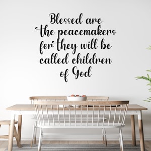May include: A white wall with a black vinyl decal that reads "Blessed are the peacemakers for they will be called children of God". The decal is above a wooden table with two white chairs and a wooden stool.
