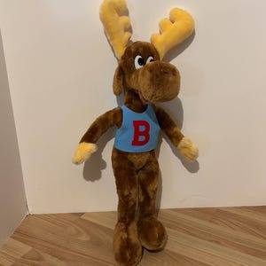 Può includere: Un alce di peluche marrone che indossa una canotta blu con una "B" rossa sul davanti. L'alce ha grandi corna gialle.