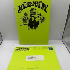 以下が含まれることがあります： ネオングリーンの雑誌と封筒。雑誌の表紙には「CREATURE FEATURE」の文字とモンスターのイラストが描かれています。封筒には同じロゴと住所情報が記載されています。