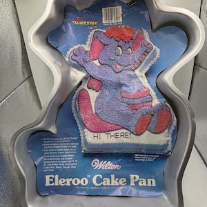 Könnte beinhalten: Eine silberne Wilton Eleroo Kuchenform mit einem Comic-Elefanten und dem Text "Hi There!" und "Wuzzles" auf der Verpackung. Die Form ist für einen 2-stöckigen Kuchen ausgelegt.