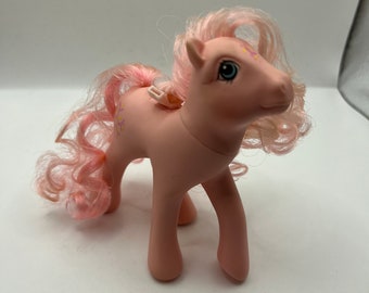 May include: A pink My Little Pony toy with a pink and white curly mane and tail. The pony has blue eyes and a pink and white flower design on its body.