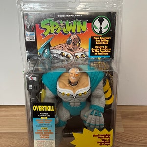 Op de afbeelding: Een Spawn Overkill action figure met een blauw, grijs en wit kleurenschema. De figuur is geposeerd met zijn rechterarm gestrekt en zijn linkerhand gebald. De figuur is verpakt in een plastic blisterverpakking met een Spawn-comic. De verpakking bevat ook de tekst "From America's Best Selling Comic Book" en "Du Livre de Bandes Dessinées le Plus Populaire en Amérique".