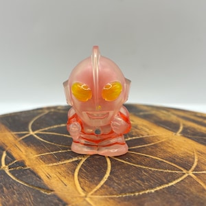 Puede incluir: Una figura translúcida rosa de un personaje de dibujos animados con ojos amarillos y rayas rojas. La figura está en posición de pie con los puños cerrados. La figura está sobre una superficie de madera con un patrón geométrico.